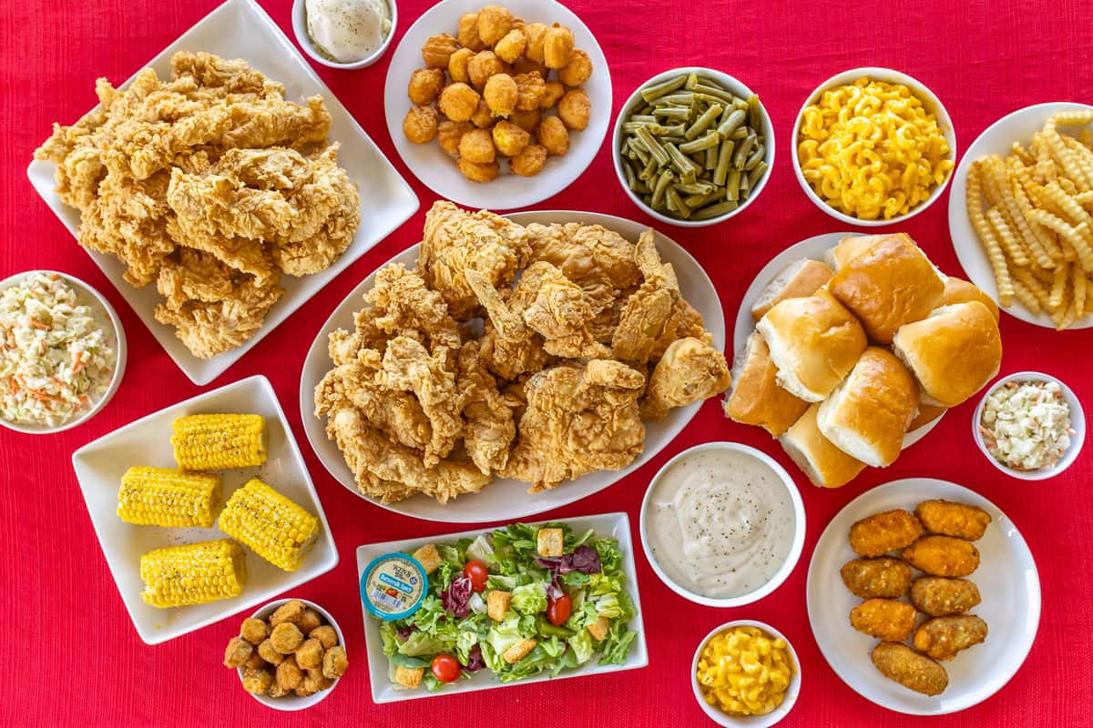 Family-style Southern comfort food spread featuring crispy fried chicken, chicken tenders, golden dinner rolls, crinkle-cut fries, mac and cheese, coleslaw, corn on the cob, green beans, fried okra, hush puppies, stuffed jalapeños, a fresh garden salad with ranch, and creamy white gravy, all served on a red tablecloth for a homestyle dining experience.