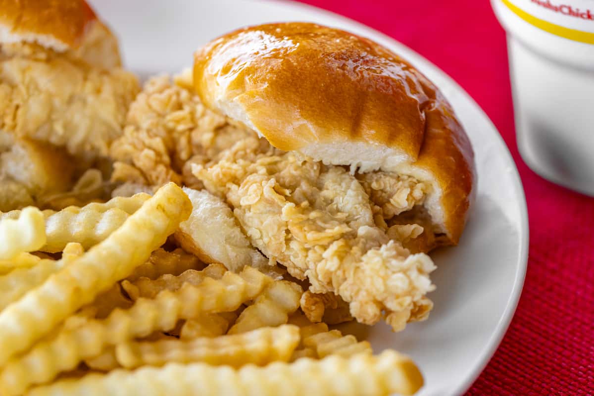 Close-up of a crispy chicken tender sandwich on a soft, glossy dinner roll, served with a side of golden crinkle-cut fries on a white plate over a red tablecloth.