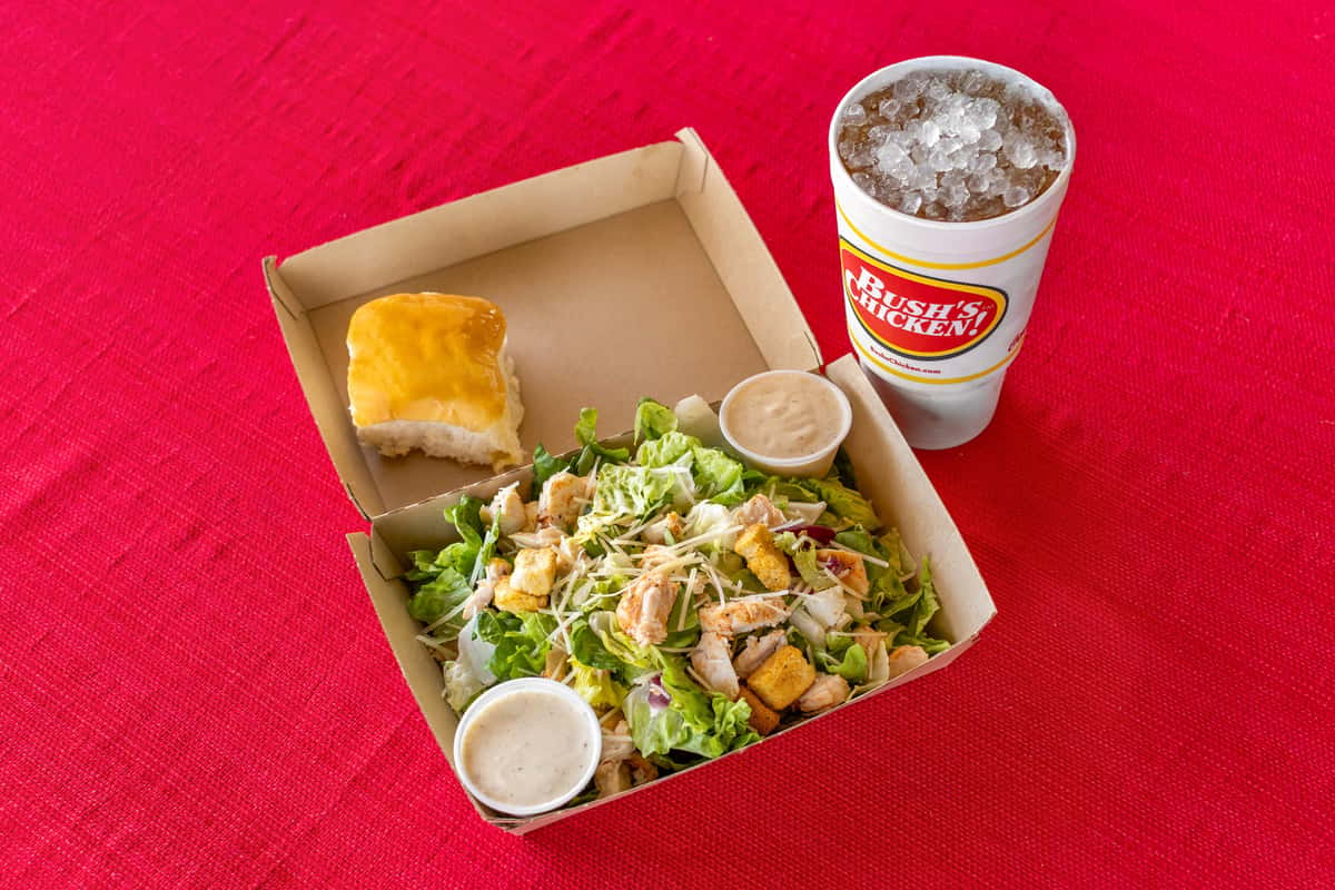 Fresh grilled chicken salad topped with shredded cheese and croutons, served with ranch dressing, a warm dinner roll, and a large iced drink from Bush's Chicken on a bright red tablecloth.