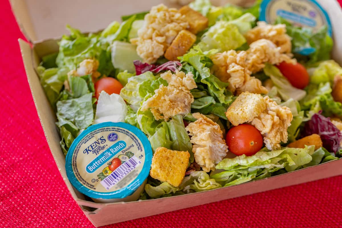 Crispy chicken salad in a takeout box featuring chopped romaine and iceberg lettuce, cherry tomatoes, golden croutons, and bite-sized fried chicken pieces, served with Ken's Buttermilk Ranch dressing on a red textured tablecloth.