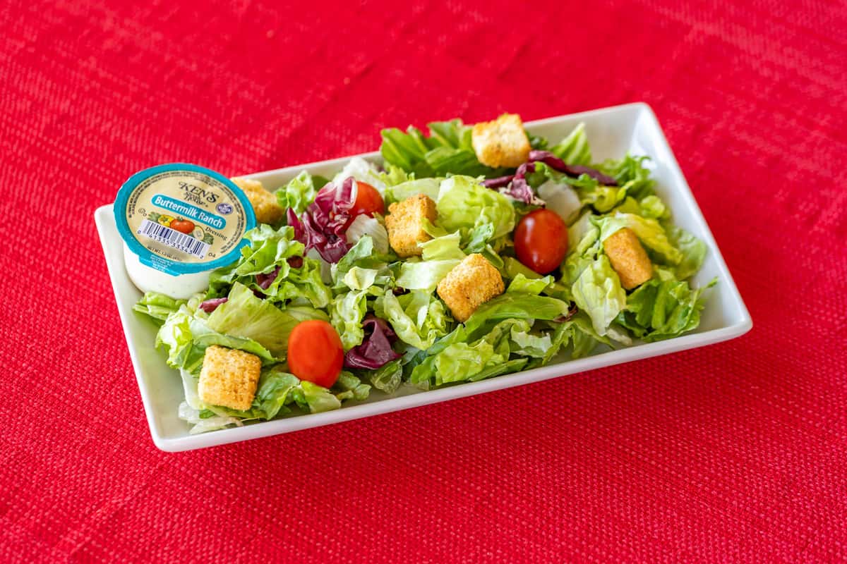 Fresh garden side salad with crisp romaine and iceberg lettuce, grape tomatoes, and crunchy croutons, served with a Ken's Buttermilk Ranch dressing cup on a rectangular white plate over a red textured tablecloth.