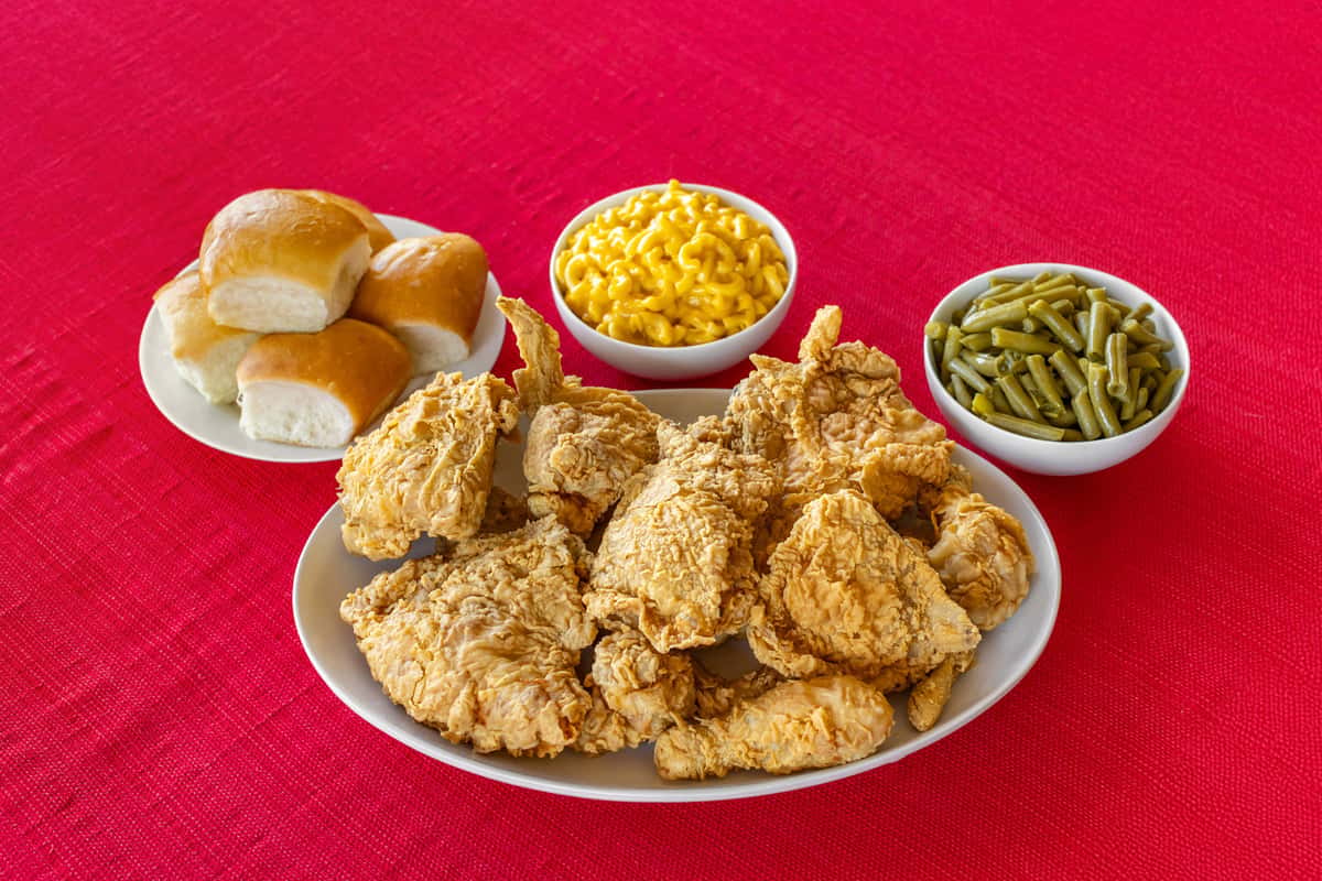 Southern fried chicken platter accompanied by buttery dinner rolls, mac and cheese, and green beans, all served on a red cloth for a comforting family meal presentation.