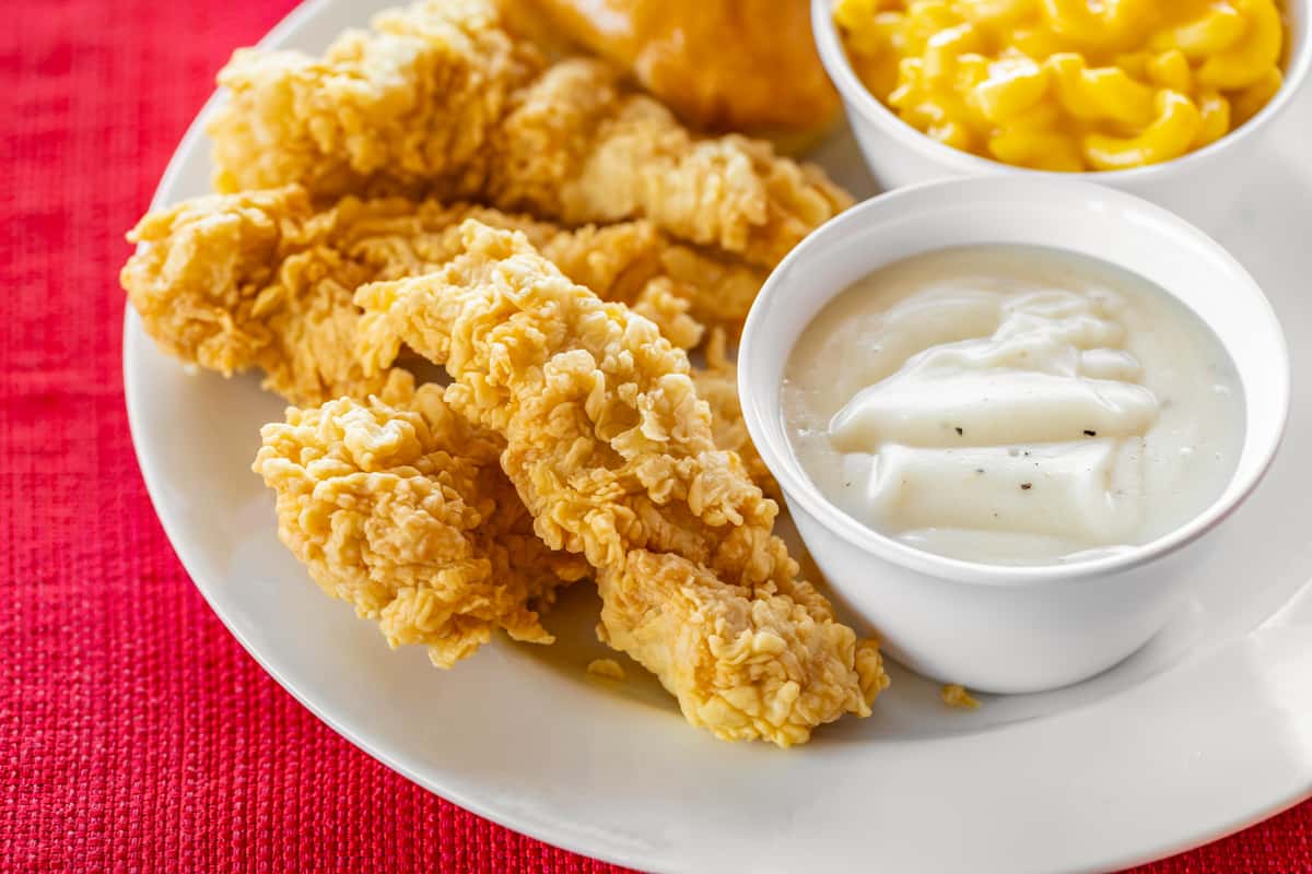 Southern-style chicken tender meal featuring crispy golden tenders, creamy peppered white gravy, mac and cheese, and a soft dinner roll, all plated on a white dish over a red woven tablecloth.