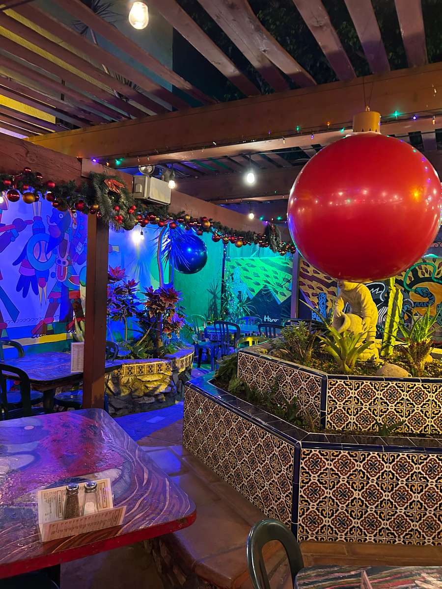 Colorful Mexican restaurant patio with festive string lights, tiled planters, murals, and a large red holiday ornament centerpiece.