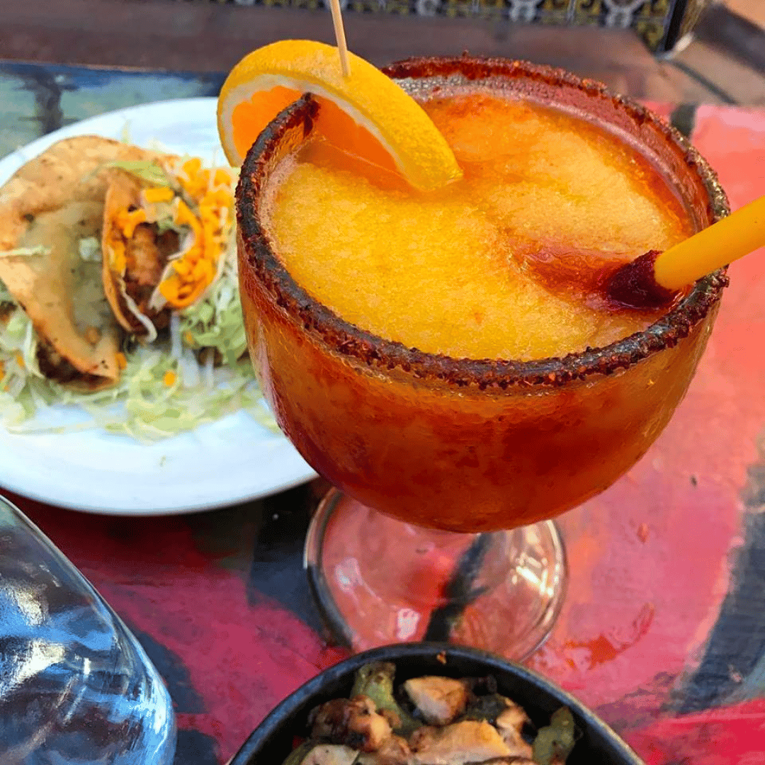 Frozen mango chamoy margarita with chili powder rim, garnished with an orange slice, with tacos and toppings in the background.