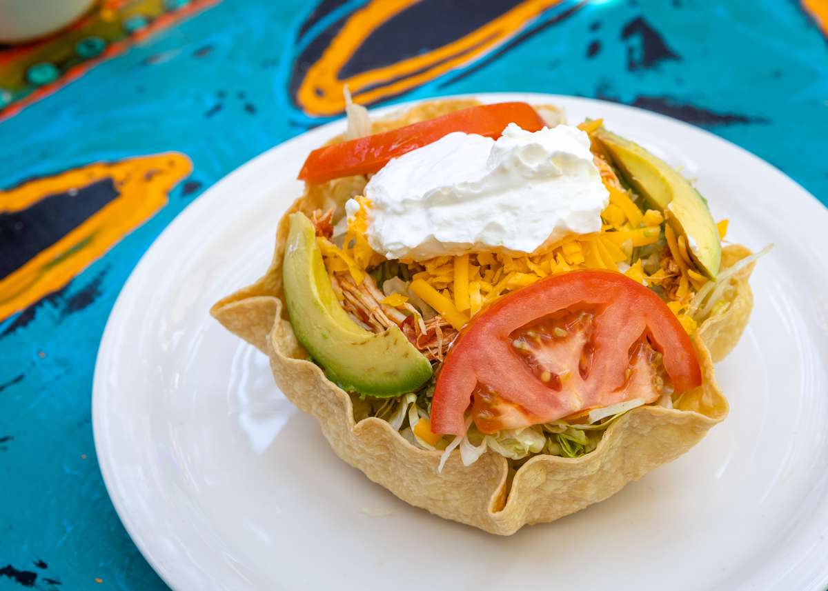 Taco salad in a crispy tortilla bowl topped with shredded cheese, avocado slices, tomato, and sour cream on a white plate.