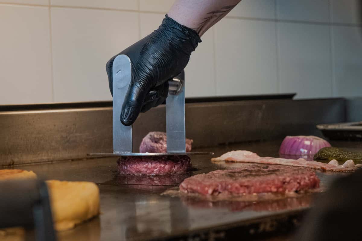 Close-up of a chef cooking a burger on a grill using a metal press, indoors.