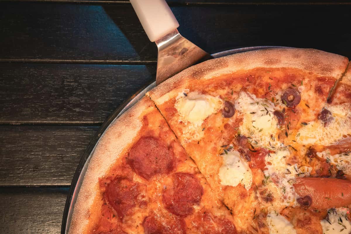Half pepperoni and half cheese pizza with olives on a metal tray, one slice being lifted with a white-handled spatula.