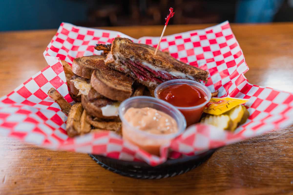 A stacked grilled sandwich loaded with smoky, savory flavor, served with crispy seasoned fries, house dipping sauce, and pickle slices on the side. Comfort food done the JAD way.