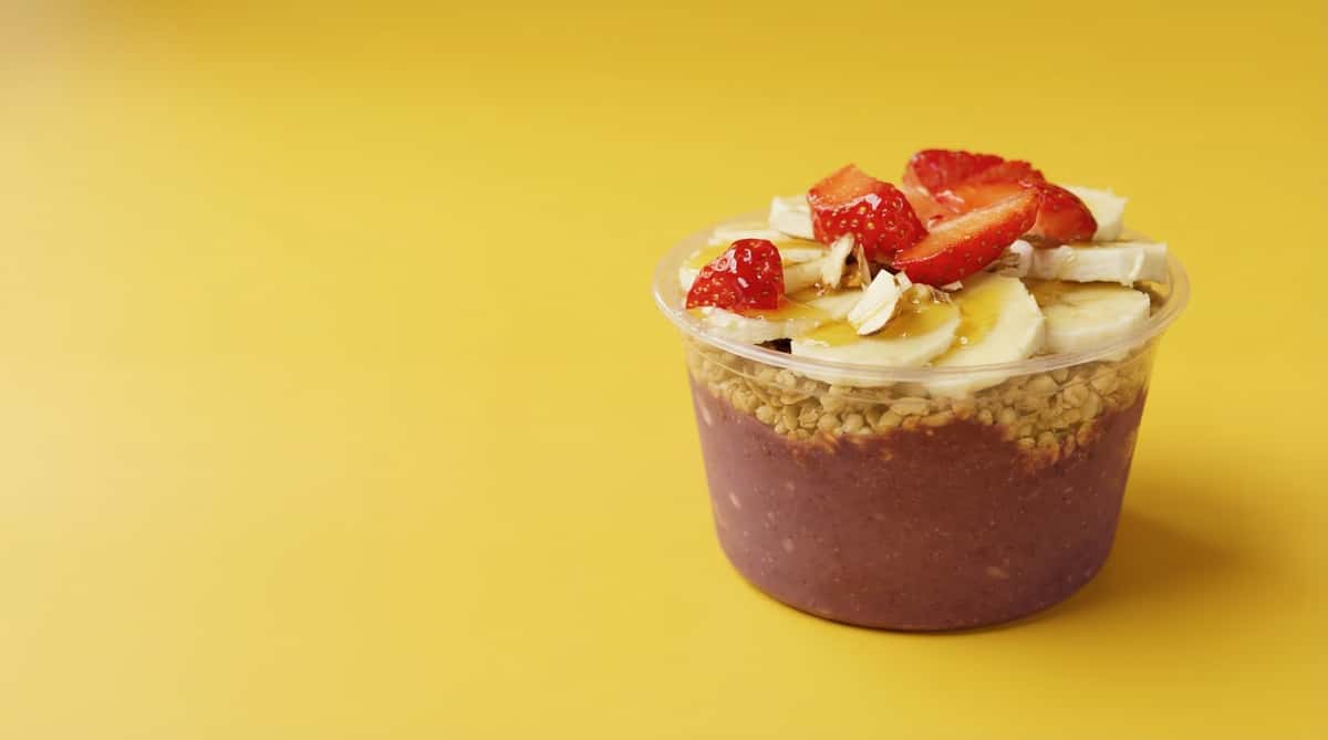 Healthy smoothie bowl with banana slices, strawberries, blueberries, granola, and chocolate base in a white bowl on a vibrant yellow surface