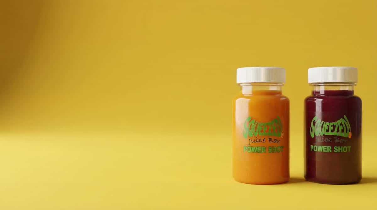 Two cold-pressed juice shots in small bottles, orange and deep red blends labeled power shots on a clean yellow background