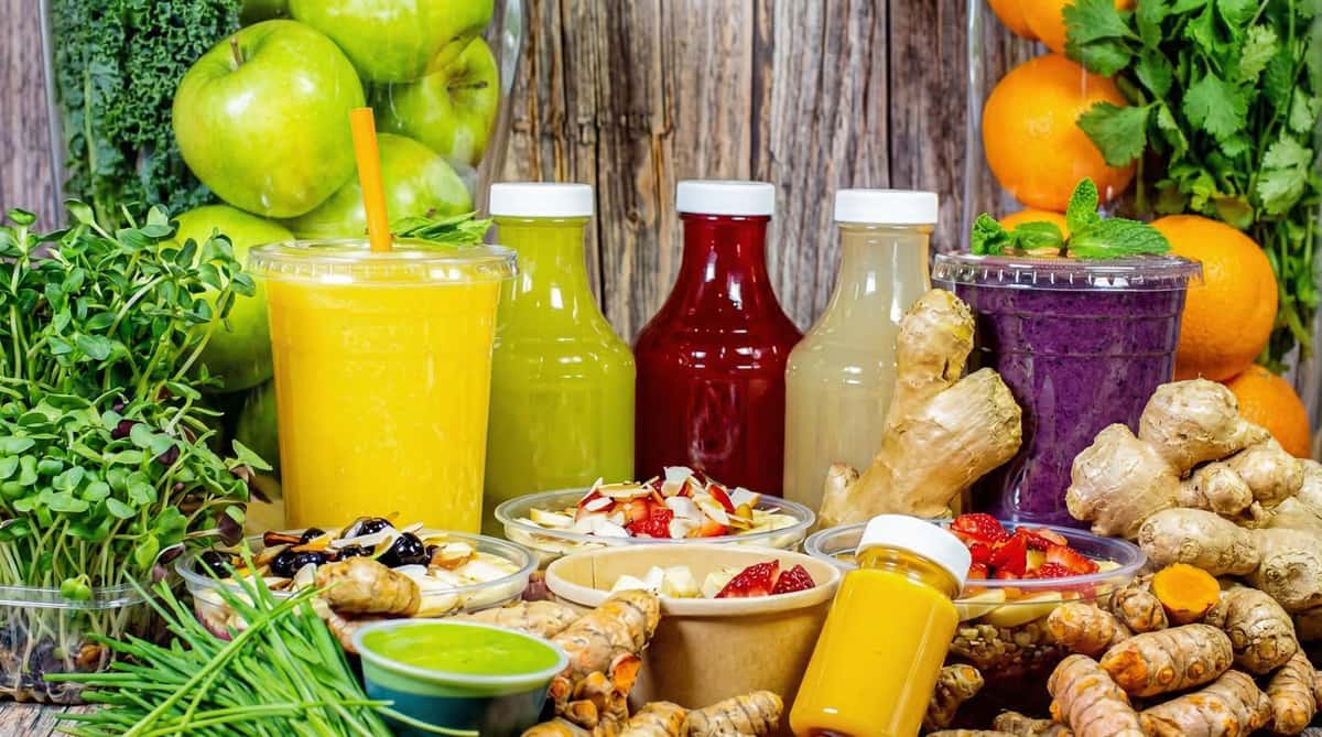 Colorful juice bar display with cold-pressed juices, smoothies, acai bowls, fresh ginger, apples, oranges, and leafy greens on a rustic wooden table