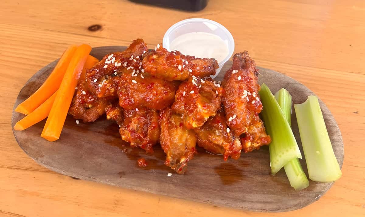 A wooden platter featuring spicy glazed chicken wings served with celery sticks, carrots, and a cup of blue cheese or ranch dip.