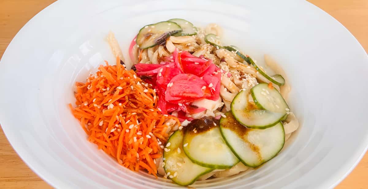 A white bowl filled with noodles, shredded carrots, cucumber slices, and pickled ginger or radish.