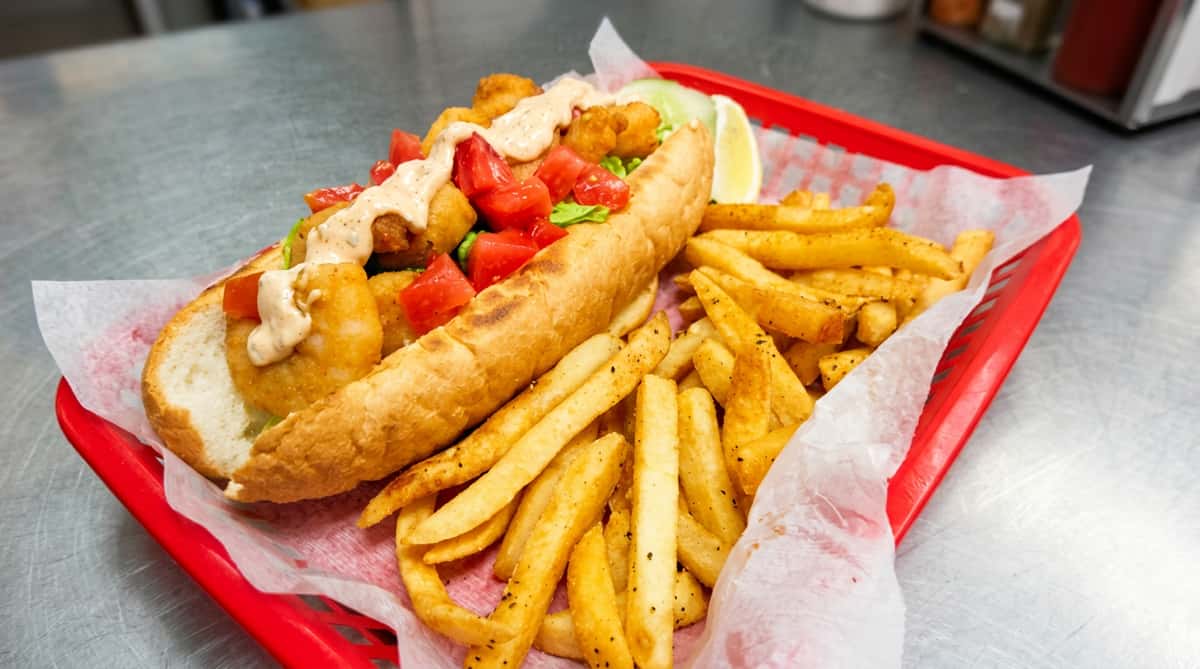 Fried shrimp po'boy with lettuce, diced tomatoes, and sauce, served with seasoned fries and lime wedge in a red basket.