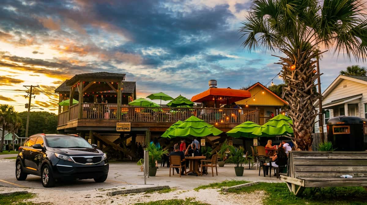 Restaurant building with two levels, outdoor seating under green and orange umbrellas, palm trees, and a parked car at sunset.