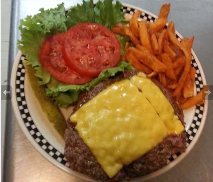 hamburger patty with a melted slice of cheese on a plate with lettuce, two tomato slices, a pickle spear and sweet potato fries