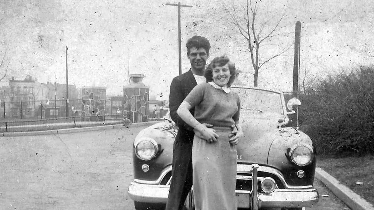 Black and White image of Maria with husband, Donald, posing in front of a car.