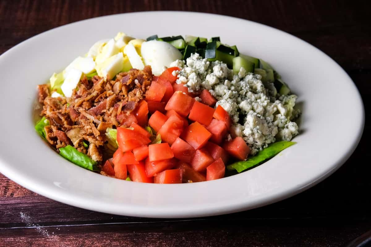 Cobb salad topped with diced tomatoes, crumbled blue cheese, chopped hard-boiled eggs, crispy bacon, and diced cucumbers over fresh lettuce, served in a white bowl.