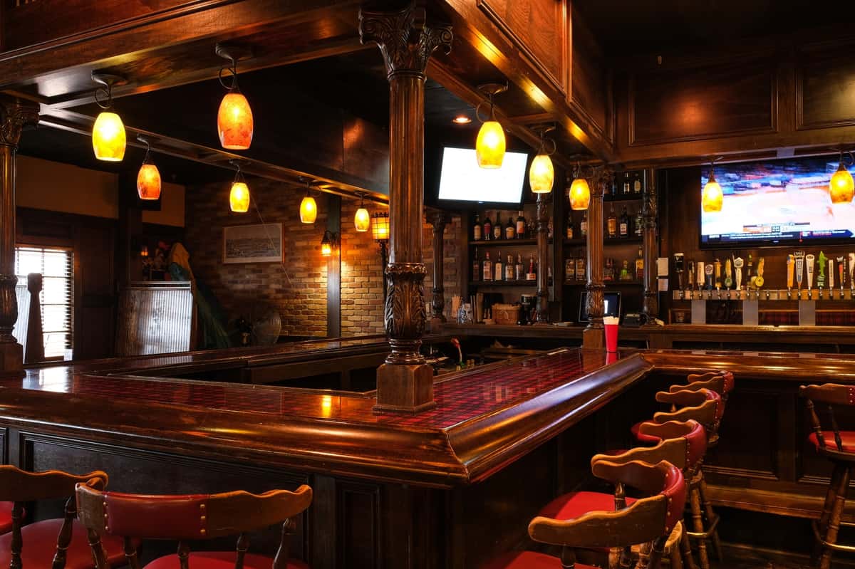 Warmly lit wood-paneled tavern interior with a polished bar, red barstools, hanging pendant lights, and TVs mounted above the bar at Ye Olde Dunwoody Tavern
