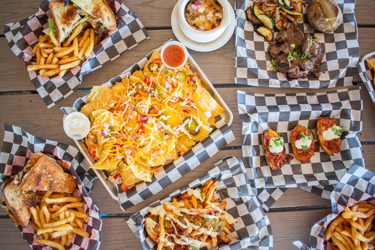 Overhead view of an assorted appetizer spread on a wooden table, including loaded fries with dipping sauces, chicken wings, and other shared plates on checkered paper liners.