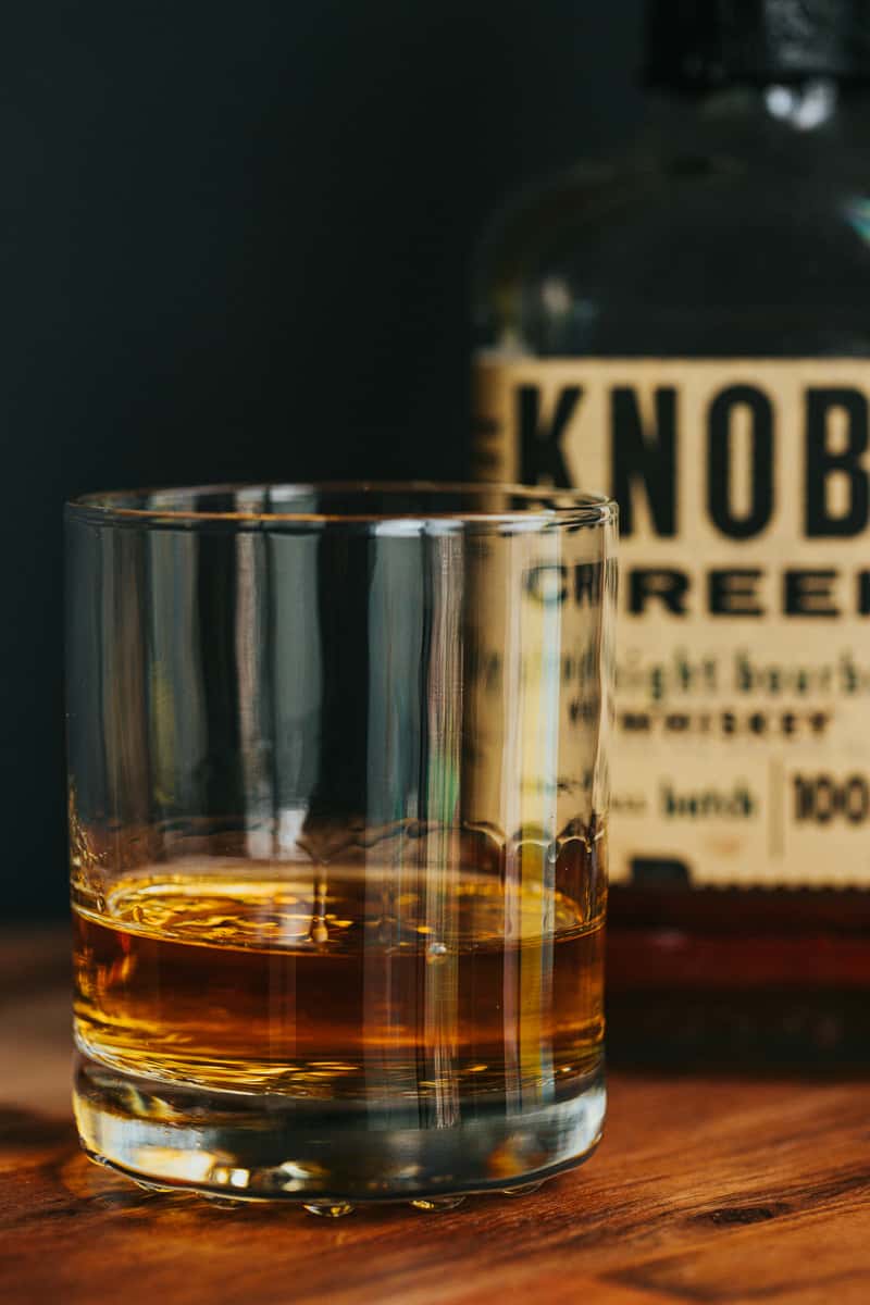 Close-up of a whiskey glass filled with Knob Creek bourbon on a rustic wooden surface, showcasing rich amber tones in a moody bar setting.