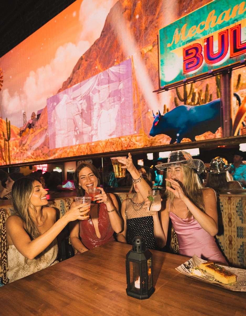 Friends raising cocktails in a lively Western-themed bar booth beneath neon bull décor, enjoying drinks and nightlife entertainment.