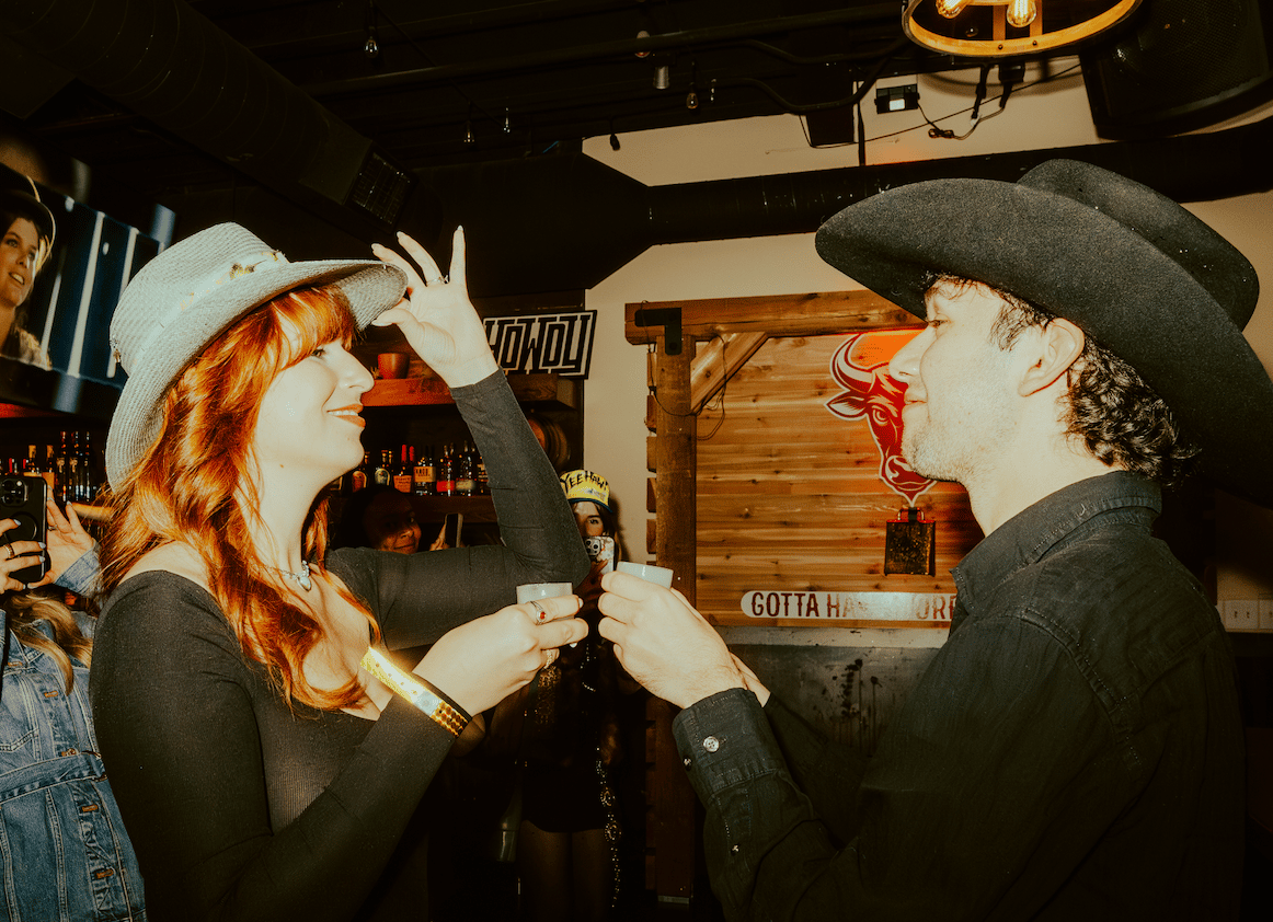 Cowboy-themed guests sharing shots and conversation inside a rustic Western bar with wood accents and neon bull signage.