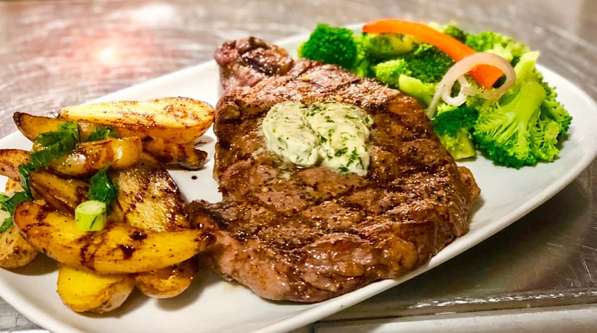 Grilled steak with herb butter, roasted potatoes, and steamed broccoli, garnished with sliced onion and red pepper.
