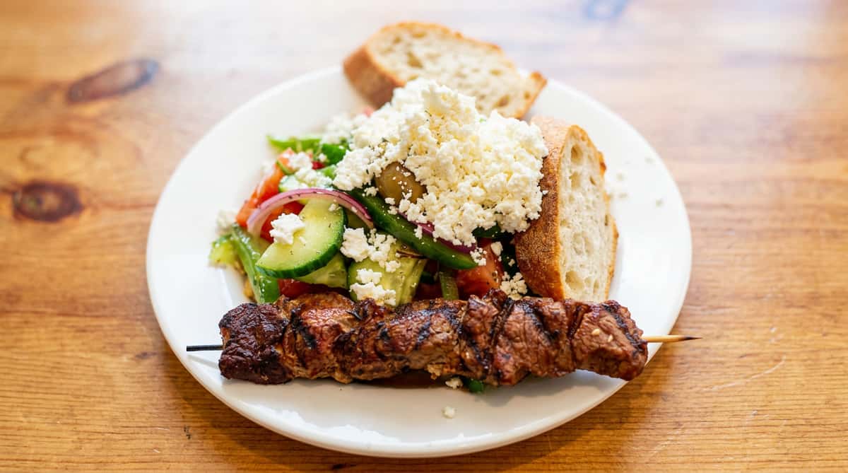 Souvlaki skewer with a side of Greek salad topped with feta cheese and two slices of bread.