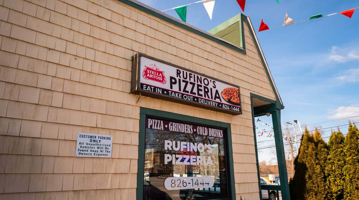 Exterior of Rufino's Pizzeria with light shingle siding, green trim, and string lights, under a clear sky.