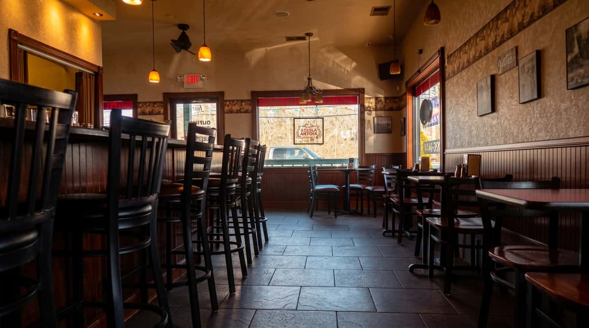 Dark restaurant interior with a bar and stools on the left, tables and chairs on the right, and large windows revealing a street outside.
