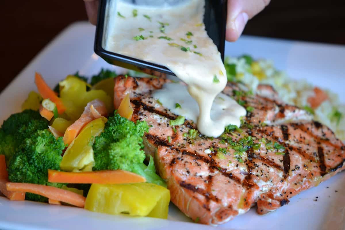 A perfectly grilled salmon fillet is topped with a creamy sauce being poured over it, enhancing its rich, flaky texture. The dish is paired with a colorful medley of fresh vegetables, adding brightness and balance. The presentation feels warm and inviting, highlighting a fresh, wholesome seafood experience.