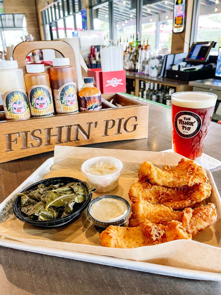 Southern-style fried catfish with collard greens, coleslaw, tartar sauce, and a pint of amber beer; BBQ sauces branded "The Fishin' Pig" in the background—authentic barbecue and soul food pairing.