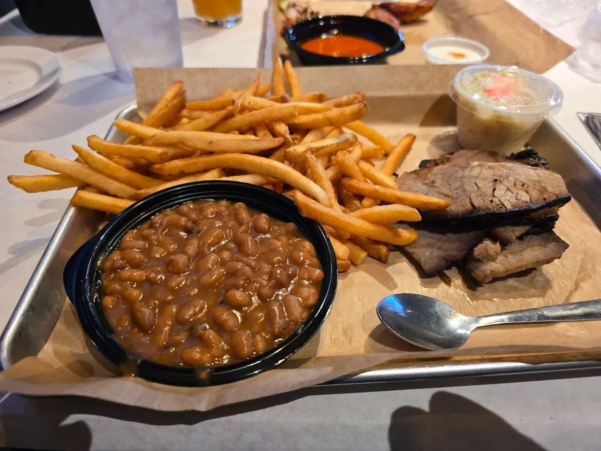 BBQ platter featuring tender smoked brisket slices, crispy golden French fries, sweet baked beans, and a side of creamy coleslaw — a classic American barbecue comfort meal served on a tray.