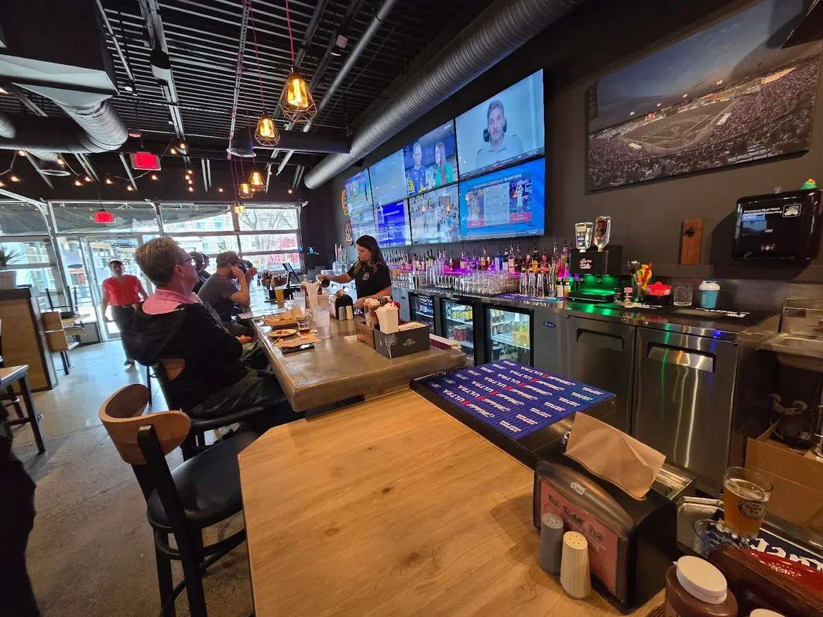 Modern sports bar interior with flat-screen TVs showing live games, a fully stocked bar with colorful lighting, and guests enjoying food and drinks in a lively atmosphere.