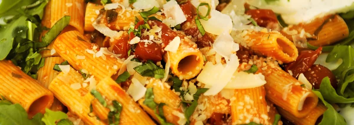 A bowl of rigatoni pasta tossed in a hearty tomato-based meat sauce, topped with shaved parmesan cheese and fresh herbs.