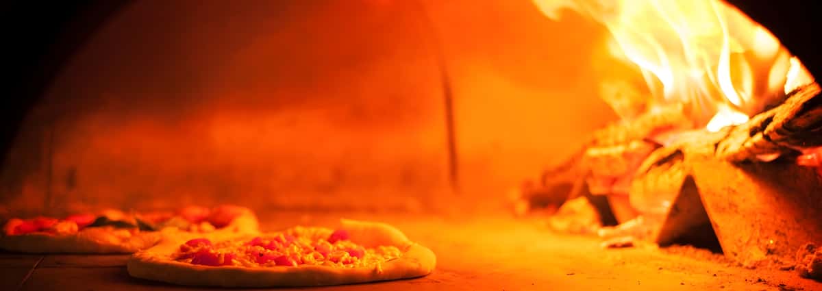A view inside a hot brick pizza oven, illuminated by bright orange flames in the background, with a pizza cooking on the stone surface.