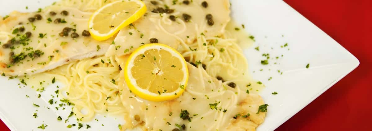 A close-up of a pasta dish featuring cutlets in a creamy lemon butter sauce, garnished with capers and fresh lemon slices.