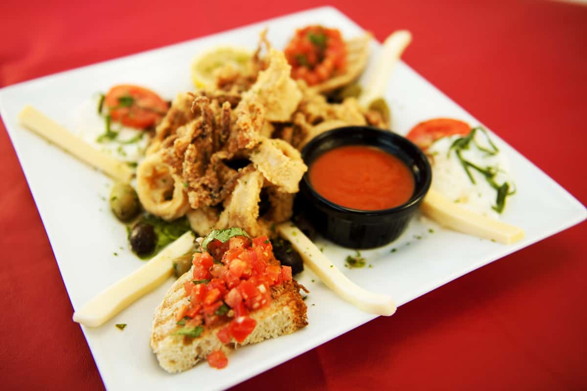 An overhead shot of a square white plate containing fried calamari, bruschetta, mozzarella sticks, and dipping sauce.