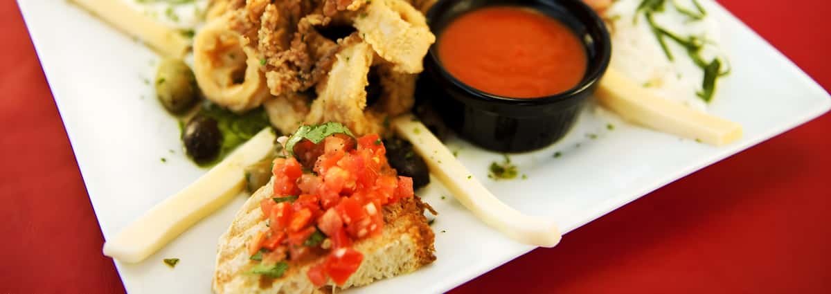 An appetizer plate featuring fried calamari rings, tomato bruschetta on toasted bread, and a ramekin of red marinara sauce.