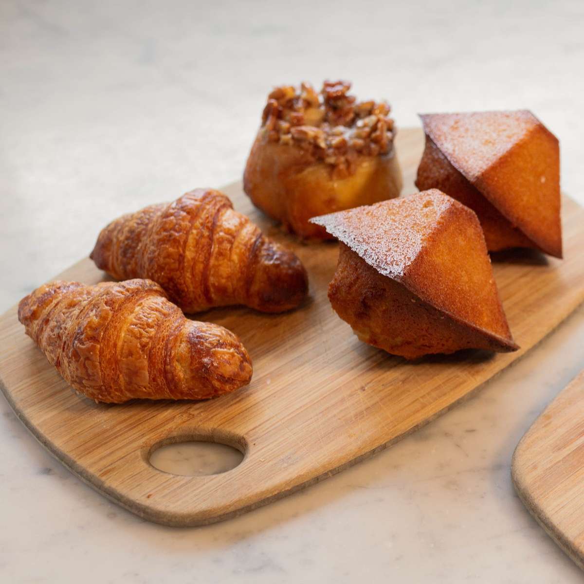 A wooden board with croissants, a nut-filled pastry, and two pyramid-shaped cakes dusted with powdered sugar.