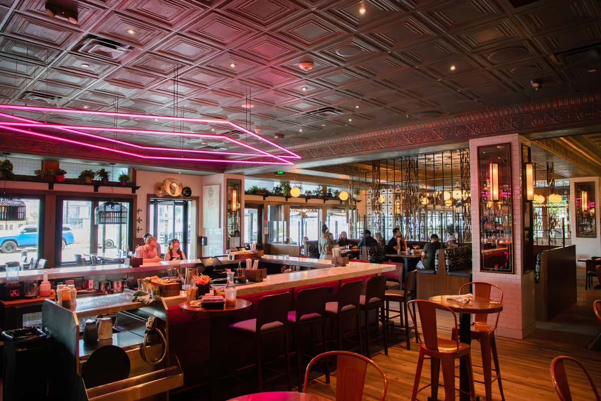 Modern cocktail bar interior featuring a neon-lit central bar, elegant mirrored accents, and contemporary dining spaces with a vibrant social atmosphere.