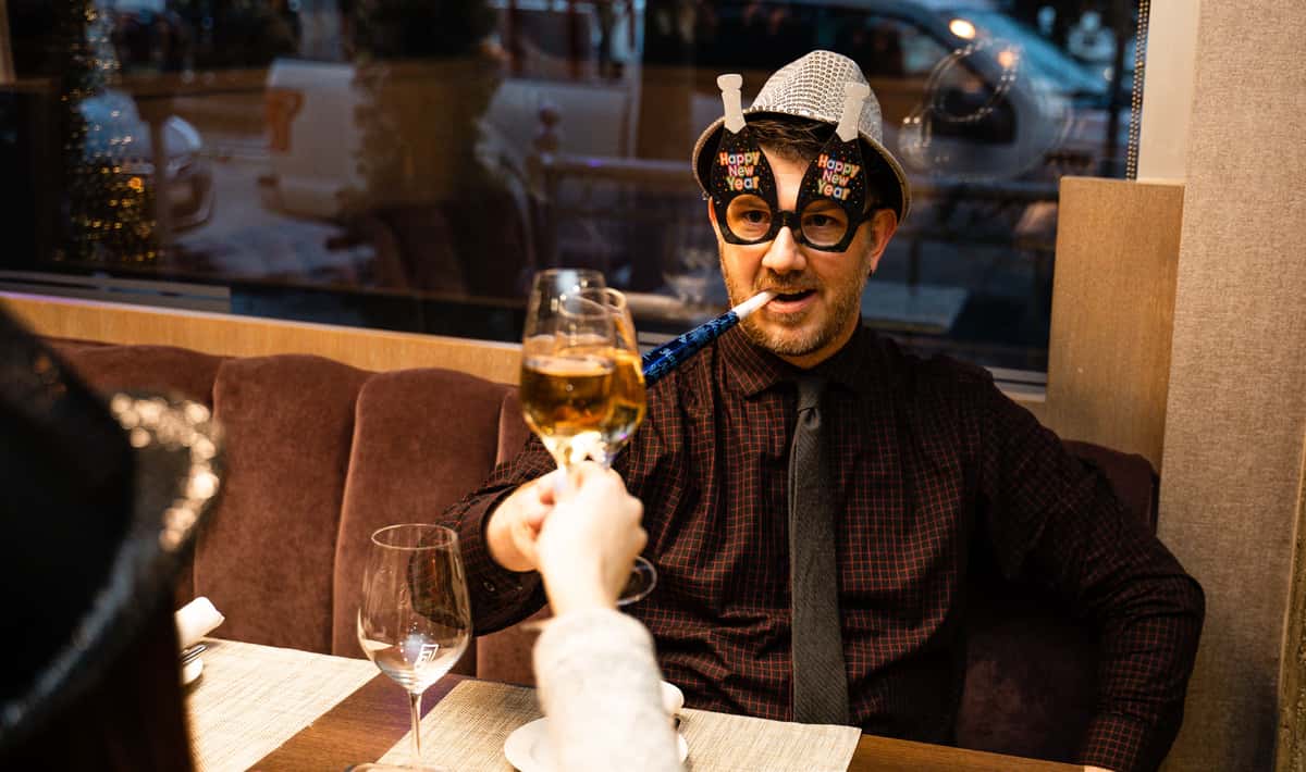 a man wearing a festive new year's hat and glasses toasts with a woman out of frame