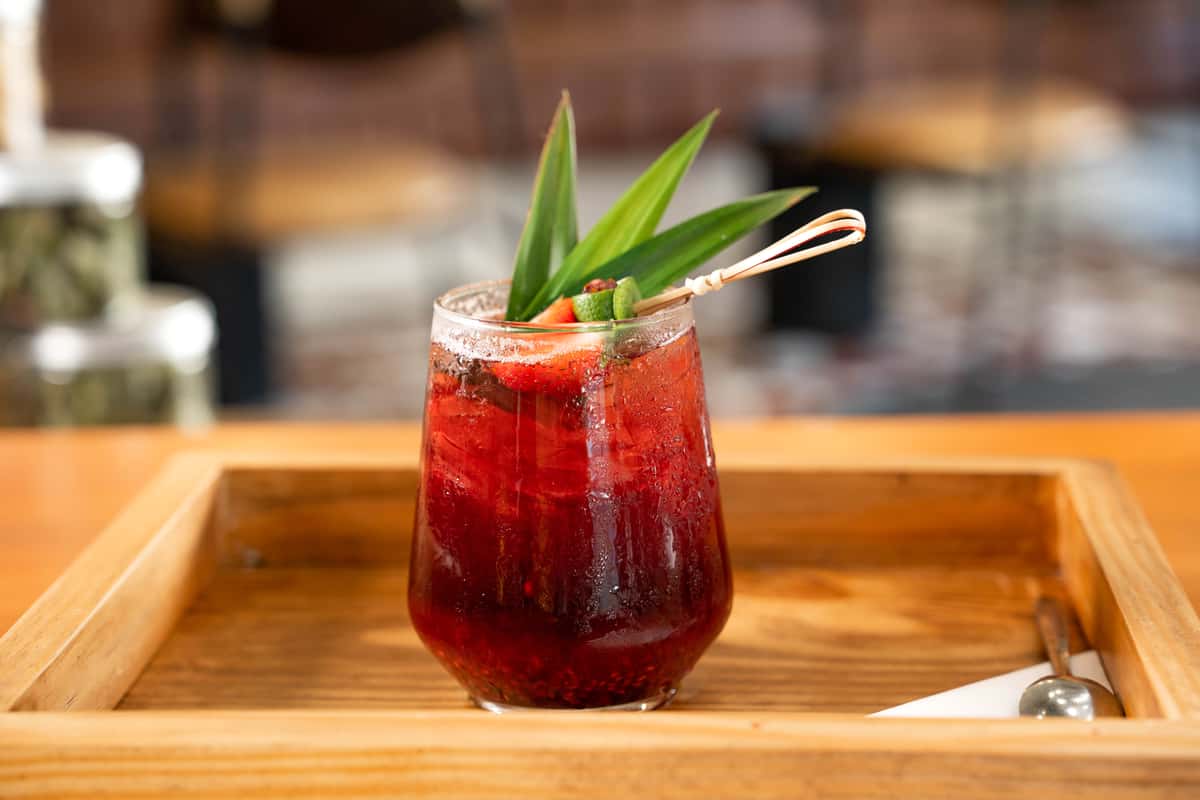 A short glass filled with a dark red iced beverage, garnished with a distinct green pandan or palm leaf.