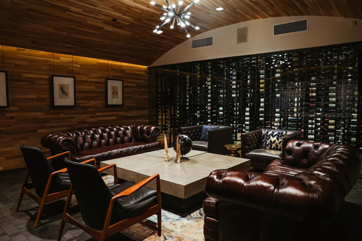 Modern lounge event space with a full-length wine rack, stylish chandelier, luxurious brown leather wingback sofas, and rich wood-panel walls.