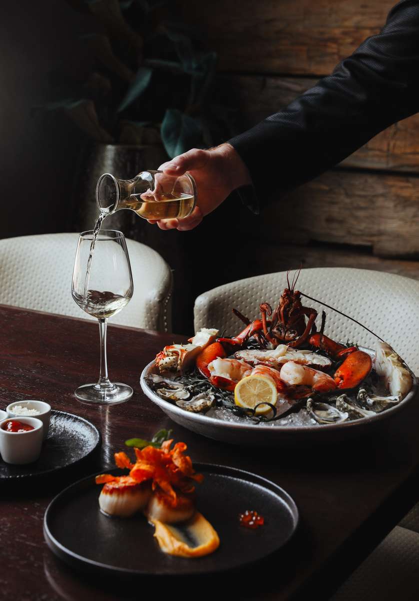 A bartender pouring wine into a glass for a patron enjoying a lavish seafood meal featuring lobster, highlighting a fine dining experience.
