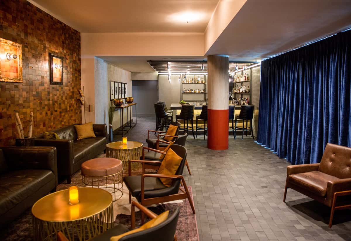 Cozy lounge area in a Nashville private event space, with leather seating, gold accent tables, ambient lighting, and a bar area with stools behind a blue velvet curtain.