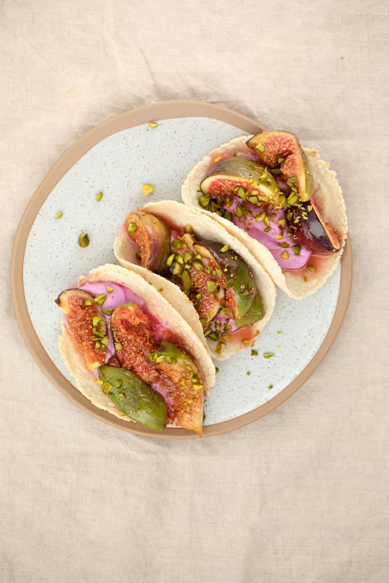 Artisan tacos with sliced figs, vibrant pink crema, and chopped pistachios on handmade corn tortillas, plated on a speckled ceramic dish over a neutral tablecloth.