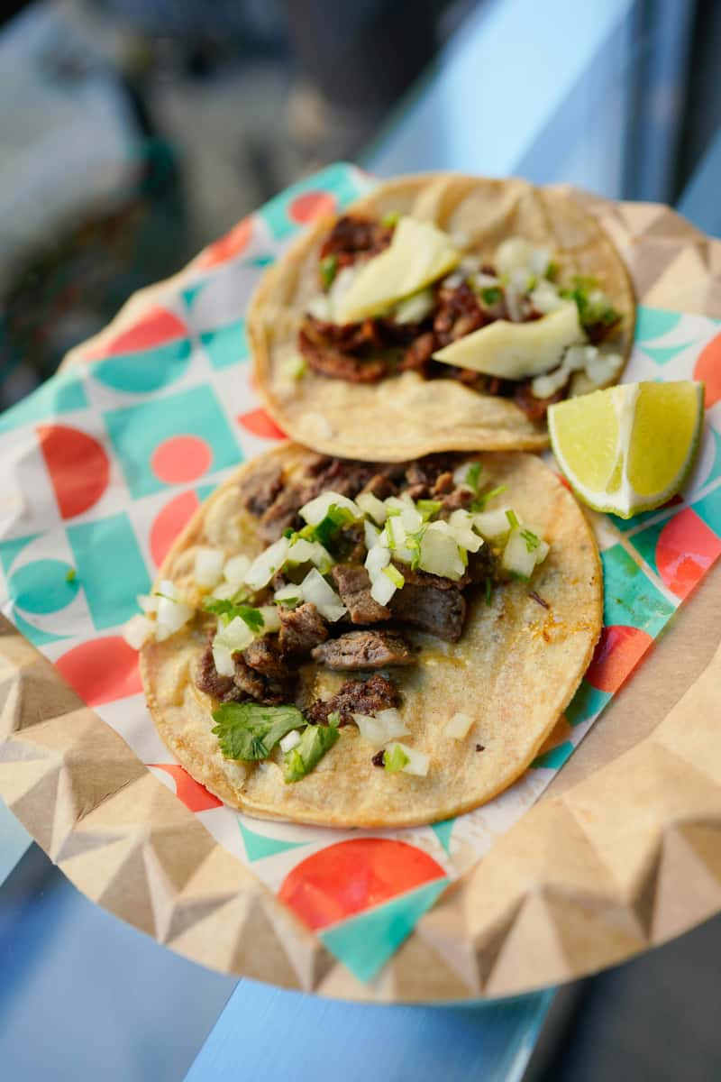 Two carne asada tacos topped with diced onions and cilantro, served with lime wedges on festive geometric wax paper in a casual street food setting.